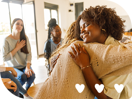 Two young women embracing during a group therapy session, while a woman in the background places her hand on her heart in admiration and approval.